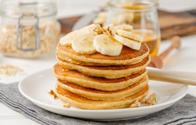 Foto da receita de Panqueca de Banana. Observa-se uma torre de panquecas altas e fofas com banana em cima e um pouco de mel escorrendo. Foto da receita de Panqueca de Banana. Observa-se uma torre de panquecas altas e fofas com banana em cima e um pouco de mel escorrendo.