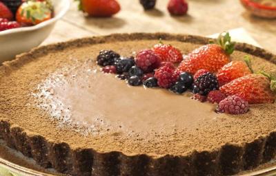 Fotografia em tons de marrom em uma mesa com uma toalha bege, um prato raso redondo grande com a torta de biscoito Nesfit com cacau e frutas vermelhas para decorar a torta, em cima do prato.