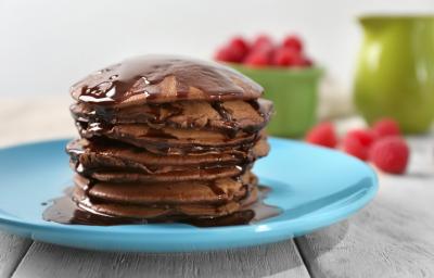 Fotografia de algumas panquecas de chocolate empilhadas em um prato azul claro, e sobre as panquecas tem uma calda de chocolate escorrendo. Ao fundo em algumas framboesas dentro de um recipiente verde, e espalhadas sobre a mesa de madeira branca. Fotografia de algumas panquecas de chocolate empilhadas em um prato azul claro, e sobre as panquecas tem uma calda de chocolate escorrendo. Ao fundo em algumas framboesas dentro de um recipiente verde, e espalhadas sobre a mesa de madeira branca.