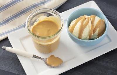 Fotografia em tons de azul e bege de uma bancada cinza, ao centro um prato retangular com o pote com uma calda de caramelo e ao lado um pote azul com duas bolas de sorvete e calda de caramelo por cima. Fotografia em tons de azul e bege de uma bancada cinza, ao centro um prato retangular com o pote com uma calda de caramelo e ao lado um pote azul com duas bolas de sorvete e calda de caramelo por cima.