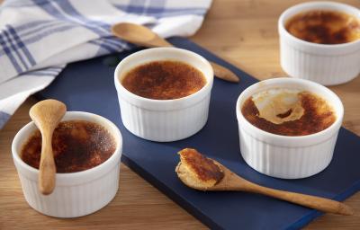Fotografia em tons de amarelo em uma bancada de madeira de cor marrom. Ao centro, uma tábua de madeira azul contendo o creme brulee. Ao lado, há algumas colheres de pau e ao fundo, um pano azul quadriculado. Fotografia em tons de amarelo em uma bancada de madeira de cor marrom. Ao centro, uma tábua de madeira azul contendo o creme brulee. Ao lado, há algumas colheres de pau e ao fundo, um pano azul quadriculado.