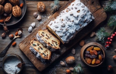 Fotografia de um bolo com frutas secas em uma tábua de madeira, polvilhado com açúcar de confeiteiro. Ao redor, nozes, amêndoas e frutas secas criam um ambiente festivo e acolhedor.