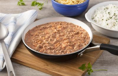 Fotografia em tons de azul em uma bancada de madeira clara com uma tábua de madeira mais escura ao centro, com uma frigideira preta grande com o strogonoff de carne moída dentro. Ao lado, um pote branco com arroz, outro azul com batata palha. Fotografia em tons de azul em uma bancada de madeira clara com uma tábua de madeira mais escura ao centro, com uma frigideira preta grande com o strogonoff de carne moída dentro. Ao lado, um pote branco com arroz, outro azul com batata palha.