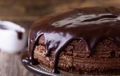 Um bolo de chocolate redondo, com textura densa e caseira, está coberto por uma espessa camada de cobertura de chocolate brilhante que escorre pelas laterais. Ele está sobre um suporte de vidro, pronto para ser servido. Um bolo de chocolate redondo, com textura densa e caseira, está coberto por uma espessa camada de cobertura de chocolate brilhante que escorre pelas laterais. Ele está sobre um suporte de vidro, pronto para ser servido.