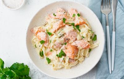 Fotografia em tons de branco, rosa, amarelo e verde de uma bancada branca vista de cima. Ao centro contém um prato redondo com macarrão, pedaços de salmão e salsa. Fotografia em tons de branco, rosa, amarelo e verde de uma bancada branca vista de cima. Ao centro contém um prato redondo com macarrão, pedaços de salmão e salsa.