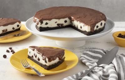 Fotografia em tons de amarelo em uma bancada cinza com um pano listrado em cinza e branco. Ao centro, um prato amarelo com uma fatia da torta de ninho com gotas de chocolate. Atrás um suporte branco com a torta inteira. Fotografia em tons de amarelo em uma bancada cinza com um pano listrado em cinza e branco. Ao centro, um prato amarelo com uma fatia da torta de ninho com gotas de chocolate. Atrás um suporte branco com a torta inteira.