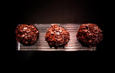 Fotografia vista de cima de três brigadeiros de tucupi reduzido recheados com cupuaçu e enrolados no chocolate amargo. Os brigadeiros estão em um apoio na cor prata, com o fundo escuro. Fotografia vista de cima de três brigadeiros de tucupi reduzido recheados com cupuaçu e enrolados no chocolate amargo. Os brigadeiros estão em um apoio na cor prata, com o fundo escuro.