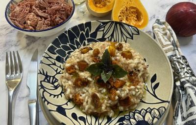 Foto da receita de risoto de carne seca e abóbora em tons de laranja servida em um prato de porcelana azul e branco. Ao lado, há talheres prateados e um guardanapo adornado, além de carne seca, abóbora e cebola ao fundo para decorar. Foto da receita de risoto de carne seca e abóbora em tons de laranja servida em um prato de porcelana azul e branco. Ao lado, há talheres prateados e um guardanapo adornado, além de carne seca, abóbora e cebola ao fundo para decorar.