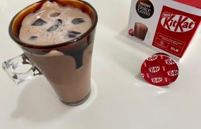 Fotografia em tons de vermelho com uma caneca de vidro ao centro. Dentro da caneca existe uma receita de Iced Mocha feita com Dolce Gusto Kitkat. Nas bordas da caneca existe uma calda de chocolate