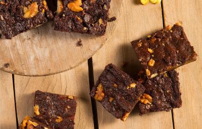 Fotografia em tons de amarelo em uma bancada de madeira com uma tábua de madeira clara com alguns brownies de chocolate em cima. Na bancada, alguns brownies espalhados e flores amarelas espalhadas na mesa para decorar. Fotografia em tons de amarelo em uma bancada de madeira com uma tábua de madeira clara com alguns brownies de chocolate em cima. Na bancada, alguns brownies espalhados e flores amarelas espalhadas na mesa para decorar.