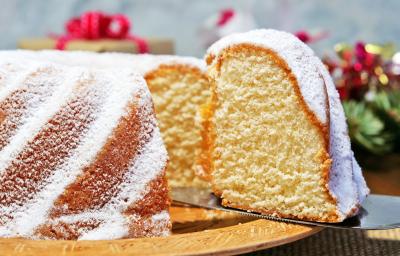 Fotografia de um bolo de massa branca de leite com um pedaço em destaque e cobertura de açúcar de confeiteiro, sobre um apoio reto de tom claro. Fotografia de um bolo de massa branca de leite com um pedaço em destaque e cobertura de açúcar de confeiteiro, sobre um apoio reto de tom claro.