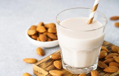 Fotografia em tons claros de um copo de vidro com um canudo dourado e branco dentro do copo, que tem uma bebida vegetal de amêndoas. O copo está sobre uma mini tábua de madeira quadrada, apoiada em uma mesa cinza clara com amêndoas espalhadas. Fotografia em tons claros de um copo de vidro com um canudo dourado e branco dentro do copo, que tem uma bebida vegetal de amêndoas. O copo está sobre uma mini tábua de madeira quadrada, apoiada em uma mesa cinza clara com amêndoas espalhadas.