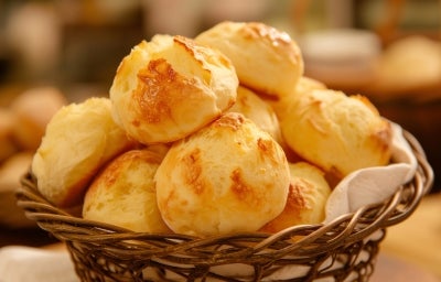 Foto da receita de Pão de Queijos. Observa-se os pães de queijo dispostos numa mesa saindo de uma cestinha de palha. Foto da receita de Pão de Queijos. Observa-se os pães de queijo dispostos numa mesa saindo de uma cestinha de palha.