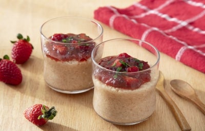Fotografia de dois potes de vidro pequenos com pudim de tapioca com geleia de morango sobre uma mesa de madeira clara. Ao redor dos recipientes têm três morangos inteiros, duas colheres de madeira pequenas e um pano vermelho e branco dobrado. Fotografia de dois potes de vidro pequenos com pudim de tapioca com geleia de morango sobre uma mesa de madeira clara. Ao redor dos recipientes têm três morangos inteiros, duas colheres de madeira pequenas e um pano vermelho e branco dobrado.