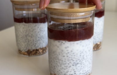 Fotografia mostra potes individuais de cheesecake, com camadas visíveis de granola, creme claro com chia e cobertura de geleia vermelha.
