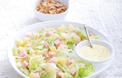 Fotografia em tons de branco e verde de uma bancada de madeira branca com paninho branco, sobre ele um recipiente redondo branco com a salada tropical e com o molho de iogurte. Ao fundo um recipiente redondo branco com o Nesfit Cereal. Fotografia em tons de branco e verde de uma bancada de madeira branca com paninho branco, sobre ele um recipiente redondo branco com a salada tropical e com o molho de iogurte. Ao fundo um recipiente redondo branco com o Nesfit Cereal.