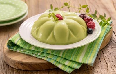 Receita de Manjar com FiberMais Suco Verde. Na foto, observa-se um manjar esverdeado sobre um prato branco que está em cima de um guardanapo em tons de verde e uma tábua oval de madeira. Frutas vermelhas e um ramo de hortelã decoram a sobremesa.
