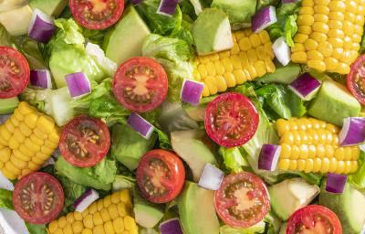 Fotografia vista de cima de uma salada mista com pequenos pedaços da espiga de milho, tomates cereja, cebola roxa, alface e chuchu. A salada está dentro de um recipiente de vidro branco, sobre uma toalha de mesa branca. Ao redor, tomates, abacate e limão. Fotografia vista de cima de uma salada mista com pequenos pedaços da espiga de milho, tomates cereja, cebola roxa, alface e chuchu. A salada está dentro de um recipiente de vidro branco, sobre uma toalha de mesa branca. Ao redor, tomates, abacate e limão.