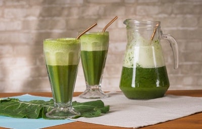 Fotografia em tons de verde com dois copos grandes ao centro. Dentro dos copos existe uma receita de suco verde feita com couve e Molico.