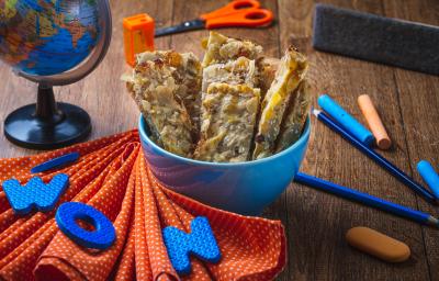 Fotografia em tons de laranja e azul em uma bancada de madeira escura, um paninho laranja e vários objetos escolares na cena. Ao centro, um potinho azul com várias barrinhas de cereal com banana e Mucilon dentro dele.