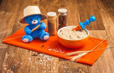 Fotografia em tons de laranja e azul de uma bancada de madeira com um paninho laranja e um pote laranja com a papinha. Na frente, grãos de arroz e cravos decorando e uma pelúcia azul com chapéu de palha.