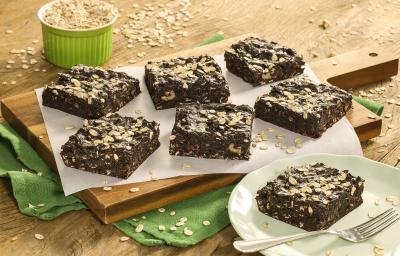 Fotografia em tons de marrom e verde de uma bancada de madeira com um paninho verde com uma tábua de madeira, sobre ela um papel manteiga com fatias de brownie. Ao lado um prato redondo verde com um brownie e um garfo. Ao fundo um pote com aveia. Fotografia em tons de marrom e verde de uma bancada de madeira com um paninho verde com uma tábua de madeira, sobre ela um papel manteiga com fatias de brownie. Ao lado um prato redondo verde com um brownie e um garfo. Ao fundo um pote com aveia.