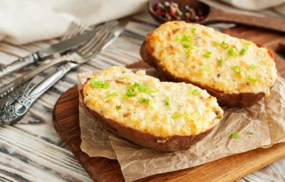 Batatas recheadas e gratinadas com queijo, finalizadas com cebolinha, sobre tábua de madeira.