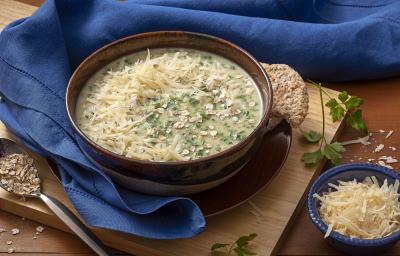 Fotografia em tons e marrom, bege e azul, com bowl marrom e bege ao centro contendo sopa com queijo e espinafre sobre prato marrom e tábua bege, ao entorno guardanapo azul, potinho com queijo e colher com aveia, tudo sobre bancada marrom. Fotografia em tons e marrom, bege e azul, com bowl marrom e bege ao centro contendo sopa com queijo e espinafre sobre prato marrom e tábua bege, ao entorno guardanapo azul, potinho com queijo e colher com aveia, tudo sobre bancada marrom.