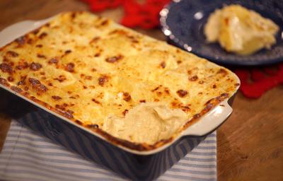 Fotografia em tons de marrom e azul de uma bancada de madeira com um paninho listrado azul e branco, sobre ele um recipiente retangular azul com o gratin. Ao fundo um paninho vermelho e um prato redondo azul com gratin.