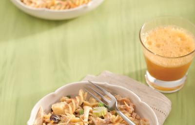 Fotografia em tons de verde em uma bancada de madeira verde com um pano bege, um prato fundo bege com a salada colorida com macarrão, ao lado um copo de vidro com um suco de laranja.