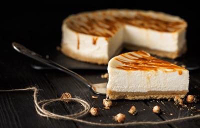Fotografia de uma fatia de cheesecake com caramelo salgado por cima de uma espátula de prata, sobre uma mesa preta. No fundo tem o resto da cheesecake sobre um apoio preto. Fotografia de uma fatia de cheesecake com caramelo salgado por cima de uma espátula de prata, sobre uma mesa preta. No fundo tem o resto da cheesecake sobre um apoio preto.