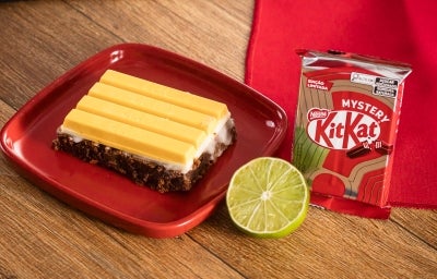 Fotografia em tons de vermelho com uma fatia de brownie ao centro. Por cima do brownie existe um glacê de limão coberto com 4 pedaços de kitkat Mystery, o kitkat de sabor misterioso de cor amarela.
