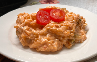 Imagem mostra uma porção de arroz cremoso em tons de dourado e salmão, com tres tomates cerejas cortados pela metade em cima. o arroz está em um prato branco, sobre uma toalha branca decorada com motivos deflores.