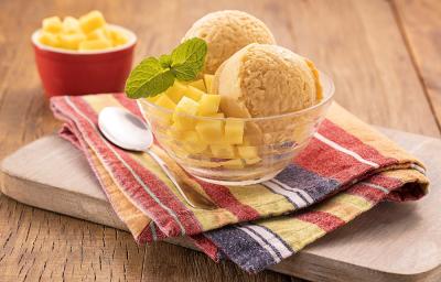 Foto da receita de sorbet de manga servida com uma folha de hortelã em uma taça de vidro sobre um pano colorido listrado. Ao lado há uma colher prateada como decoração, bem como um recipiente vermelho com mangas picadas ao fundo.