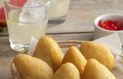 Fotografia em tons de dourado e grafite, com cestinha de vime ao centro contendo 6 coxinhas fritas, no lado direito potinho com pimentas vermelhas, ao alto dois copos com suco e gelo e um guardanapo vermelho e branco, tudo sobre bancada rústica. Fotografia em tons de dourado e grafite, com cestinha de vime ao centro contendo 6 coxinhas fritas, no lado direito potinho com pimentas vermelhas, ao alto dois copos com suco e gelo e um guardanapo vermelho e branco, tudo sobre bancada rústica.