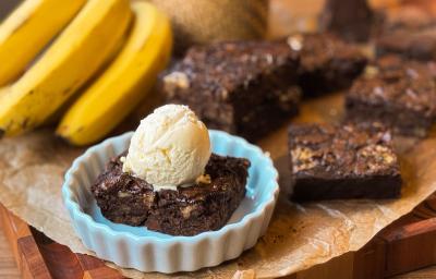 Fotografia de uma mini forma de vidro branca com um pedaço do brownie de banana com uma bola de sorvete de creme por cima. O recipiente está sobre uma folha de papel manteiga com mais pedaços do brownie e um cacho de bananas. Fotografia de uma mini forma de vidro branca com um pedaço do brownie de banana com uma bola de sorvete de creme por cima. O recipiente está sobre uma folha de papel manteiga com mais pedaços do brownie e um cacho de bananas.