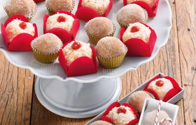 Fotografia em tons de vermelho em uma bancada de madeira com um suporte branco para bolo e vários brigadeiros de churros e com goiabada em cima dele dentro de forminhas vermelhas de docinhos.