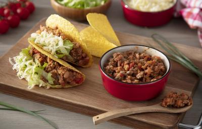 Fotografia em tons de marrom em uma bancada de madeira de cor marrom. Ao centro, há uma tábua de madeira contendo os tacos e ao lado, há uma mini panela vermelha com os recheios. Ao fundo, há tomate cereja, arroz e alface. Fotografia em tons de marrom em uma bancada de madeira de cor marrom. Ao centro, há uma tábua de madeira contendo os tacos e ao lado, há uma mini panela vermelha com os recheios. Ao fundo, há tomate cereja, arroz e alface.