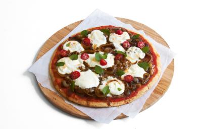 Ao centro, uma tábua de madeira marrom redonda com um pano branco e uma pizza com tomates, cebolas e iogurte por cima.