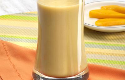 Fotografia em tons de laranja em uma bancada de madeira com uma toalha colorida listrada, um pano laranja e um copo de vidro alto com a vitamina de banana com cupuaçu. Ao fundo, um pratinho branco raso com fatias de manga. Fotografia em tons de laranja em uma bancada de madeira com uma toalha colorida listrada, um pano laranja e um copo de vidro alto com a vitamina de banana com cupuaçu. Ao fundo, um pratinho branco raso com fatias de manga.