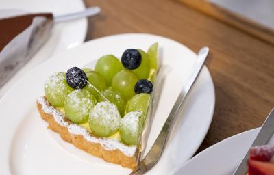 Fotografia de uma fatia de torta recheada com leite MOÇA e uvas verdes por cima, com três uvas roxas. Por cima da torta tem açúcar de confeiteiro polvilhado, e ao lado, um garfo sobre um prato branco, que está sobre uma mesa de madeira. Fotografia de uma fatia de torta recheada com leite MOÇA e uvas verdes por cima, com três uvas roxas. Por cima da torta tem açúcar de confeiteiro polvilhado, e ao lado, um garfo sobre um prato branco, que está sobre uma mesa de madeira.