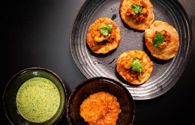 Fotografia vista de cima de um prato preto raso com quatro unidades de pani puri com grão de bico e coentro. Ao lado tem dois molhos em recipientes diferentes, um molho de iogurte e o outro, um molho de manga. Tudo está sobre uma mesa preta.