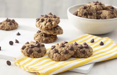 Fotografia em tons de branco e amarelo de uma bancada branca com uma tabua branca e um paninho listrado amarelo e branco sobre ele cookies. Ao fundo um recipiente redondo branco com cookies e gotas de chocolate espalhadas pela mesa.