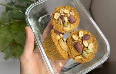 Fotografia em tons claros de uma mão segurando um recipiente de vidro pequeno e retangular. Dentro do recipiente tem três muffins de banana e aboora, decorados com castanhas.