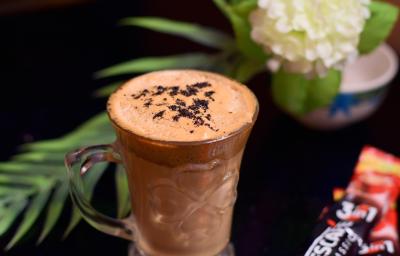 Fotografia de uma xícara de vidro com uma bebida com chocolate, Nescafé e leite. Ao lado, uma embalagem de Nescafé, e ao fundo, decoração de plantas e flores, sobre uma mesa preta. Fotografia de uma xícara de vidro com uma bebida com chocolate, Nescafé e leite. Ao lado, uma embalagem de Nescafé, e ao fundo, decoração de plantas e flores, sobre uma mesa preta.