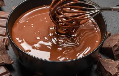 Imagem de um pequeno recipiente contendo uma calda de chocolate cremosa e derretida, em tons de marrom. Um batedor de arame mexe mistura. Ao fundo, barras de chocolate. Imagem de um pequeno recipiente contendo uma calda de chocolate cremosa e derretida, em tons de marrom. Um batedor de arame mexe mistura. Ao fundo, barras de chocolate.