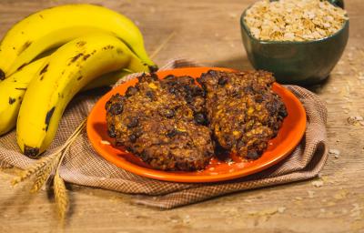 Foto da receita de Cookie Fitness de Banana e Neston, sobre um prato laranja em uma bancada de madeira decorada com mais bananas, trigo e uma caneca com cereais