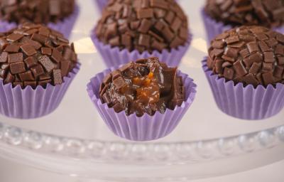 Fotografia em tons de roxo com várias forminhas de brigadeiro roxas ao centro. Dentro de cada forminha existe um brigadeiro de chocolate meio amargo recheado com um creme de caramelo salgado.