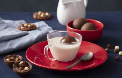 Foto de uma bancada escura. No centro há um prato vermelho com uma xícara com leite no centro e, dentro dela, há uma esfera de chocolate boiando. Ao lado do prato há mais algumas esferas fechadas, uma aberta, alguns cookies e cereal espalhado