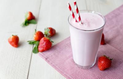 Fotografia de uma vitamina de morango em um copo de vidro com dois canudos nas cores branco em vermelho, o copo está apoiado sobre uma toalha vermelha com bolinhas brancas. Ao lado e encima, morangos inteiros, que estão sobre uma mesa de cor clara. Fotografia de uma vitamina de morango em um copo de vidro com dois canudos nas cores branco em vermelho, o copo está apoiado sobre uma toalha vermelha com bolinhas brancas. Ao lado e encima, morangos inteiros, que estão sobre uma mesa de cor clara.